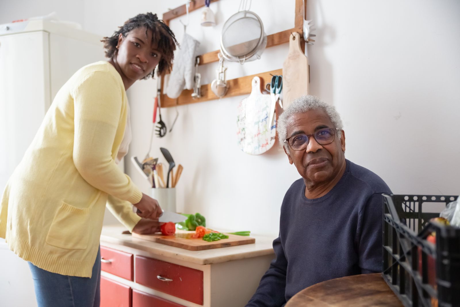 A caregiver chopping fresh vegetables while an older man sits nearby in his kitchen