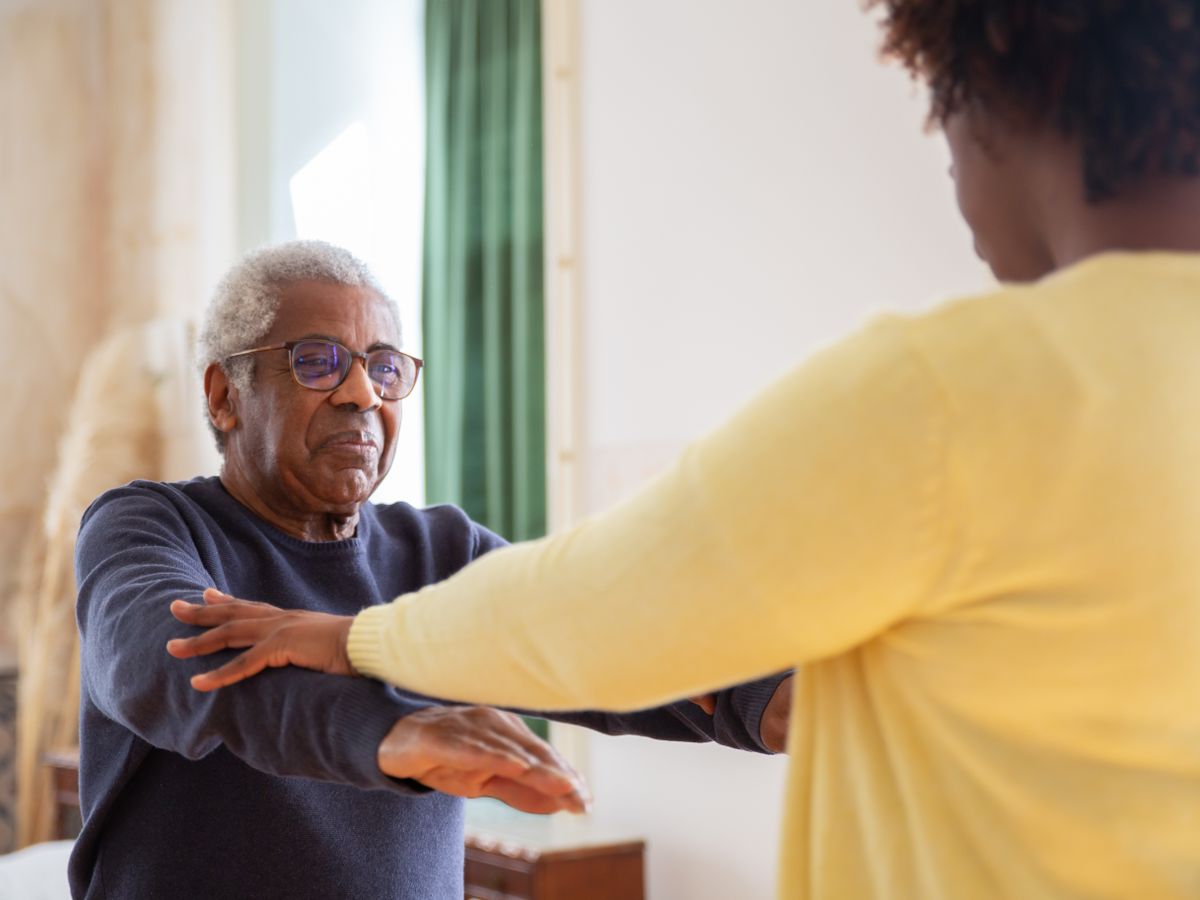A caregiver gently supporting an older man with a seated stretch at home